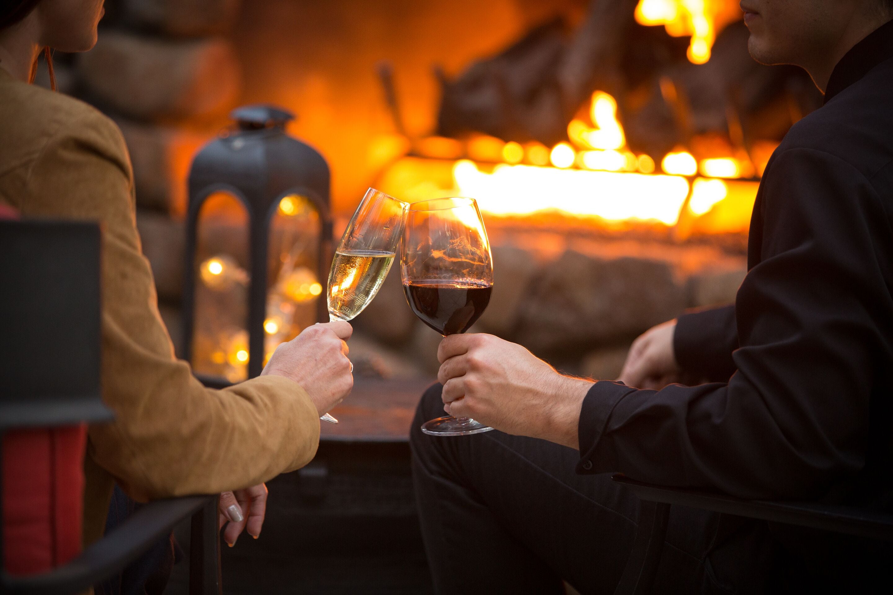 Two people toasting with wine glasses at a table with a warm, glowing background, possibly a fireplace.