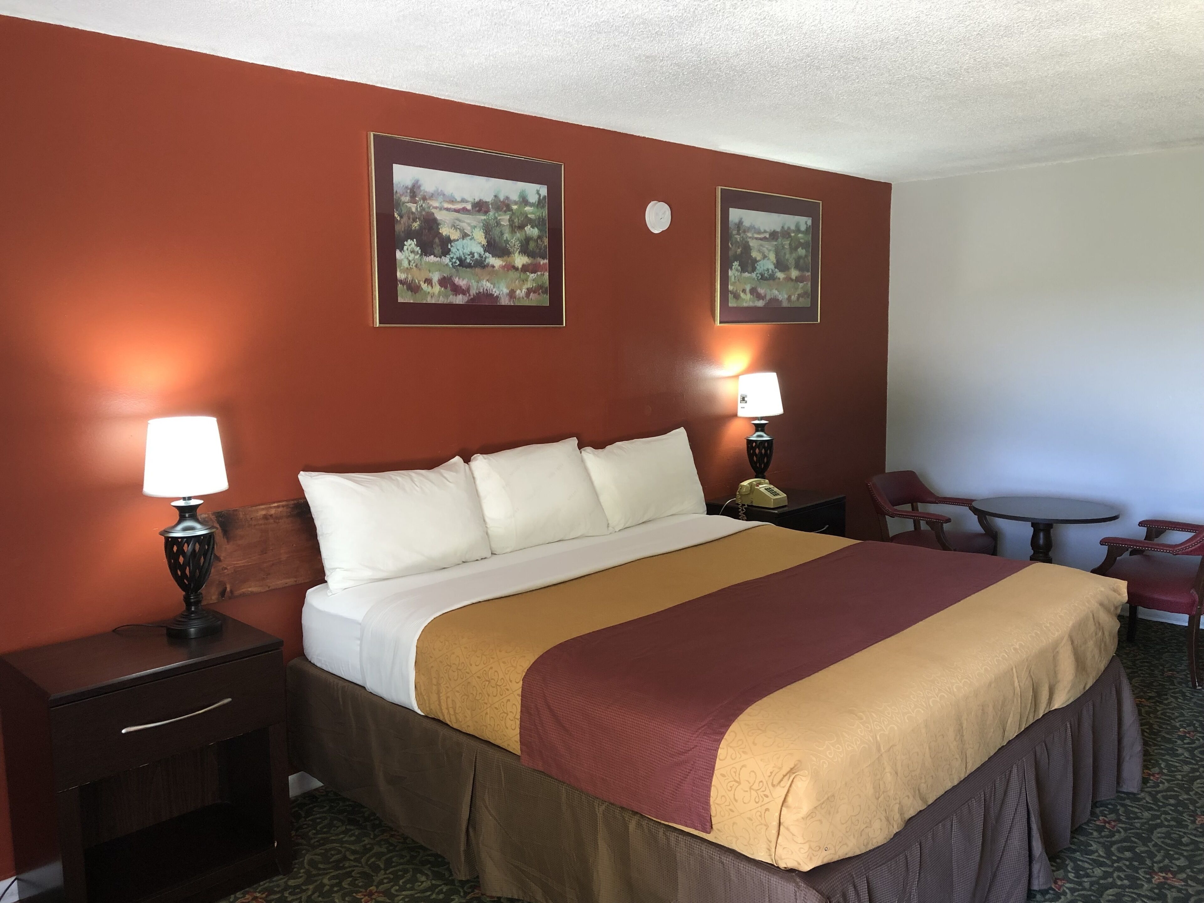Ein Hotelzimmer mit einem großen Bett, zwei Nachttischen mit Lampen, einem kleinen runden Tisch mit Stühlen und zwei gerahmten Landschaftsbildern an den Wänden.