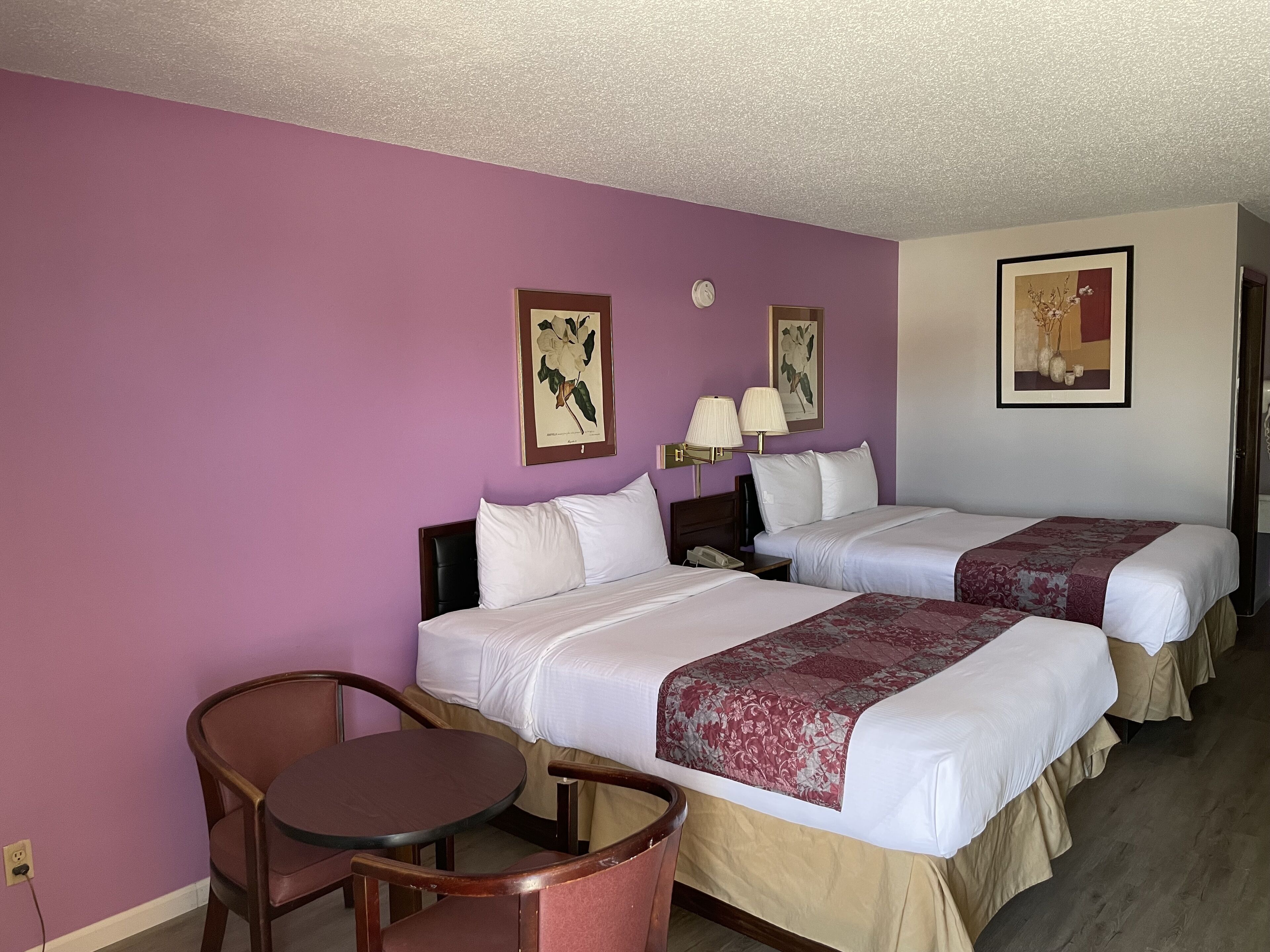 A hotel room with two beds, a small table, and chairs. There are wall-mounted lamps and framed pictures on the walls.