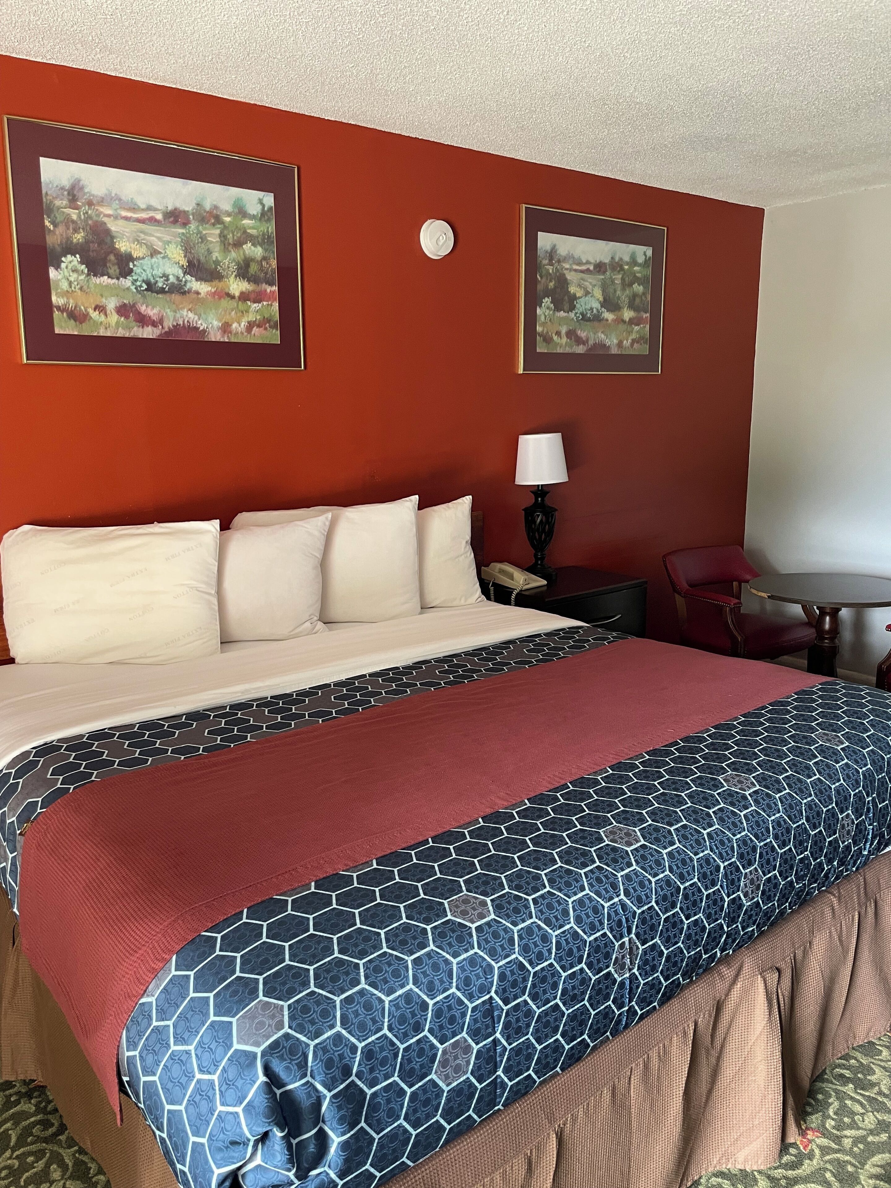 Habitación de hotel con una cama grande, un escritorio con lámpara, una silla y dos cuadros con paisajes en la pared.
