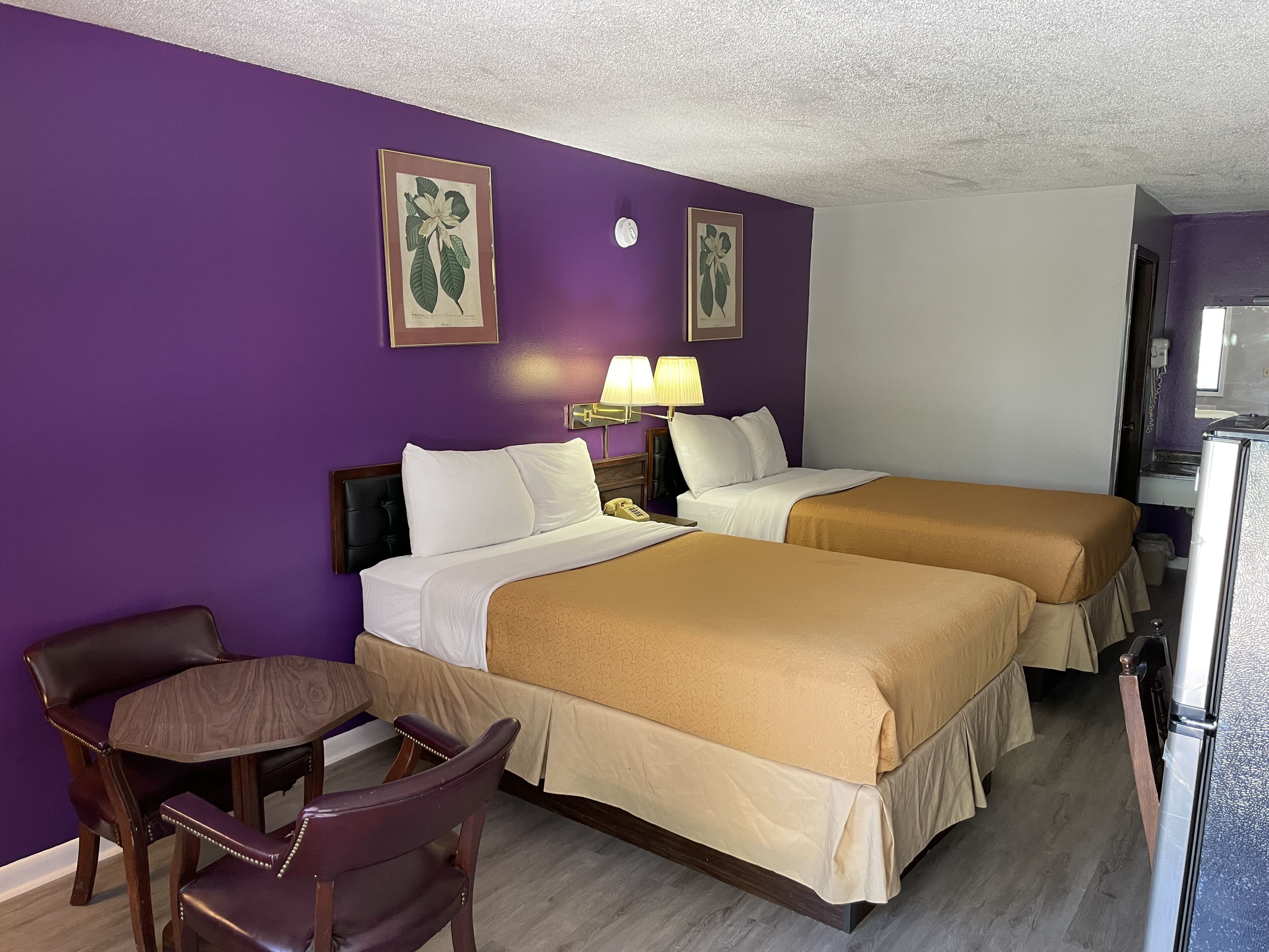 A hotel room with two beds, a desk, and a chair. There are two wall-mounted lamps and a framed botanical print on the wall.