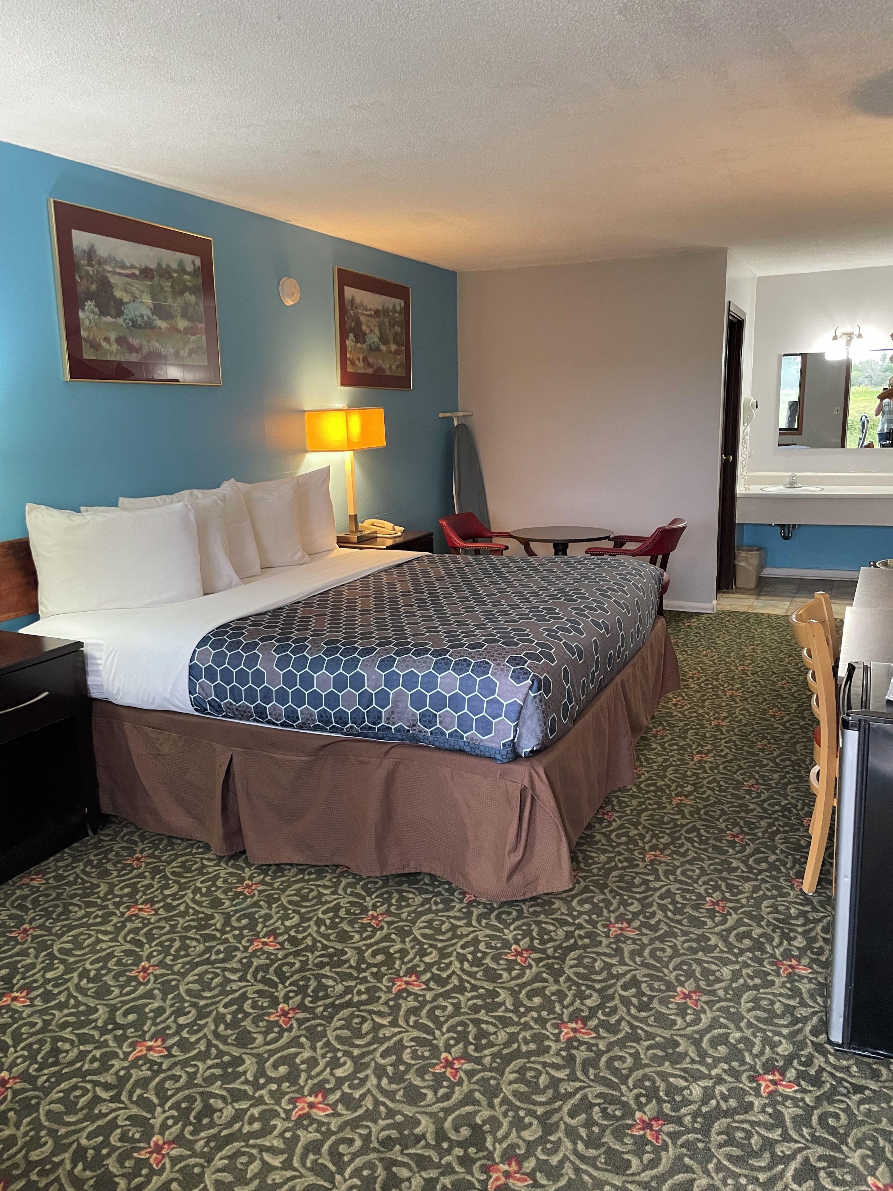 Habitación de hotel con cama, escritorio, silla, lámpara, espejo y piso alfombrado.