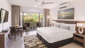 A hotel room with a bed, a desk, a chair, a TV, a ceiling fan, and a large window with a view of greenery.