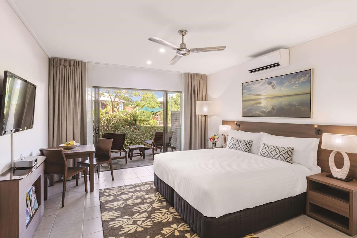 A hotel room with a bed, a desk, a chair, a TV, a ceiling fan, and a large window with a view of greenery.