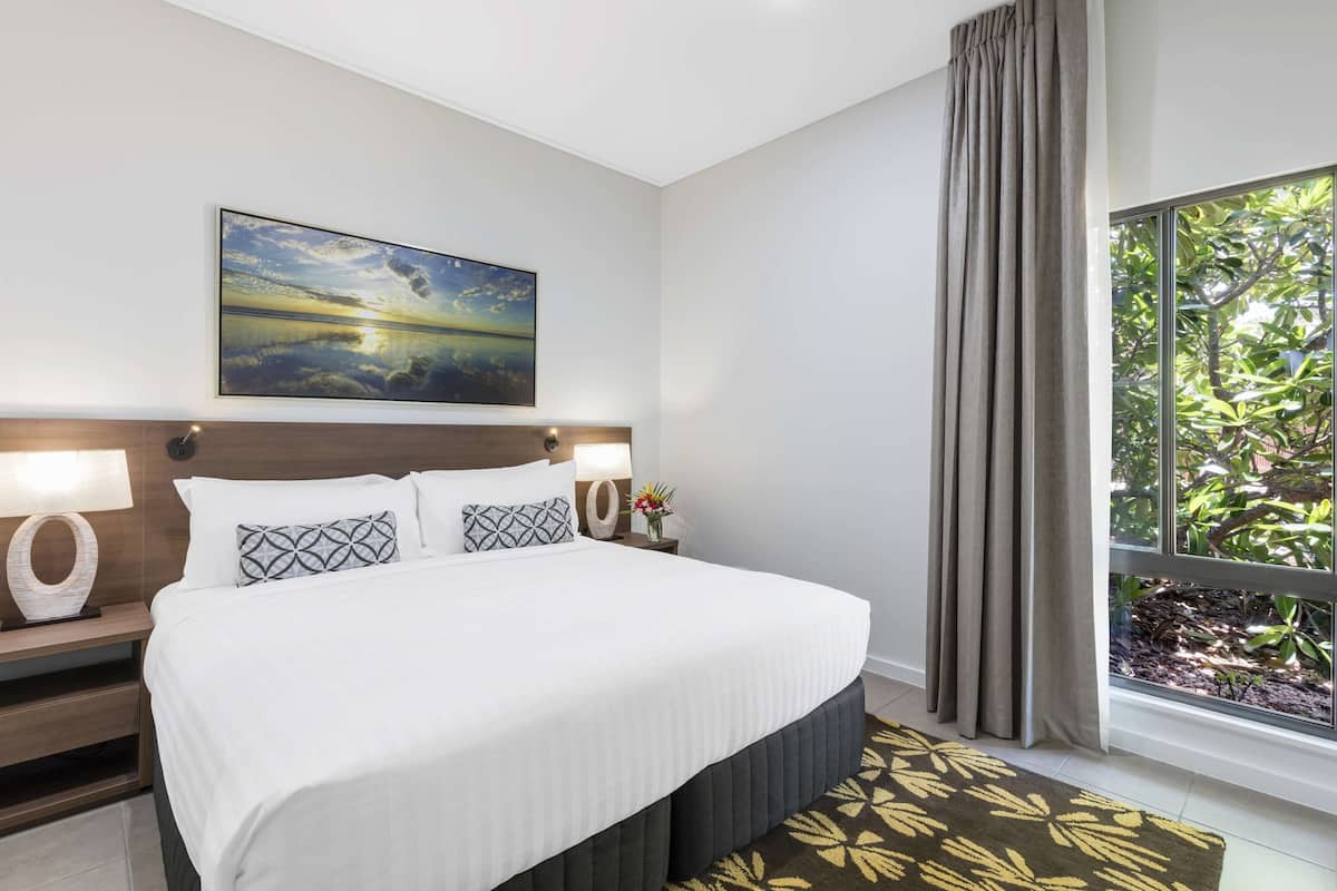 A hotel room with a bed, a nightstand, a lamp, a painting, a window with curtains, and a view of greenery outside.