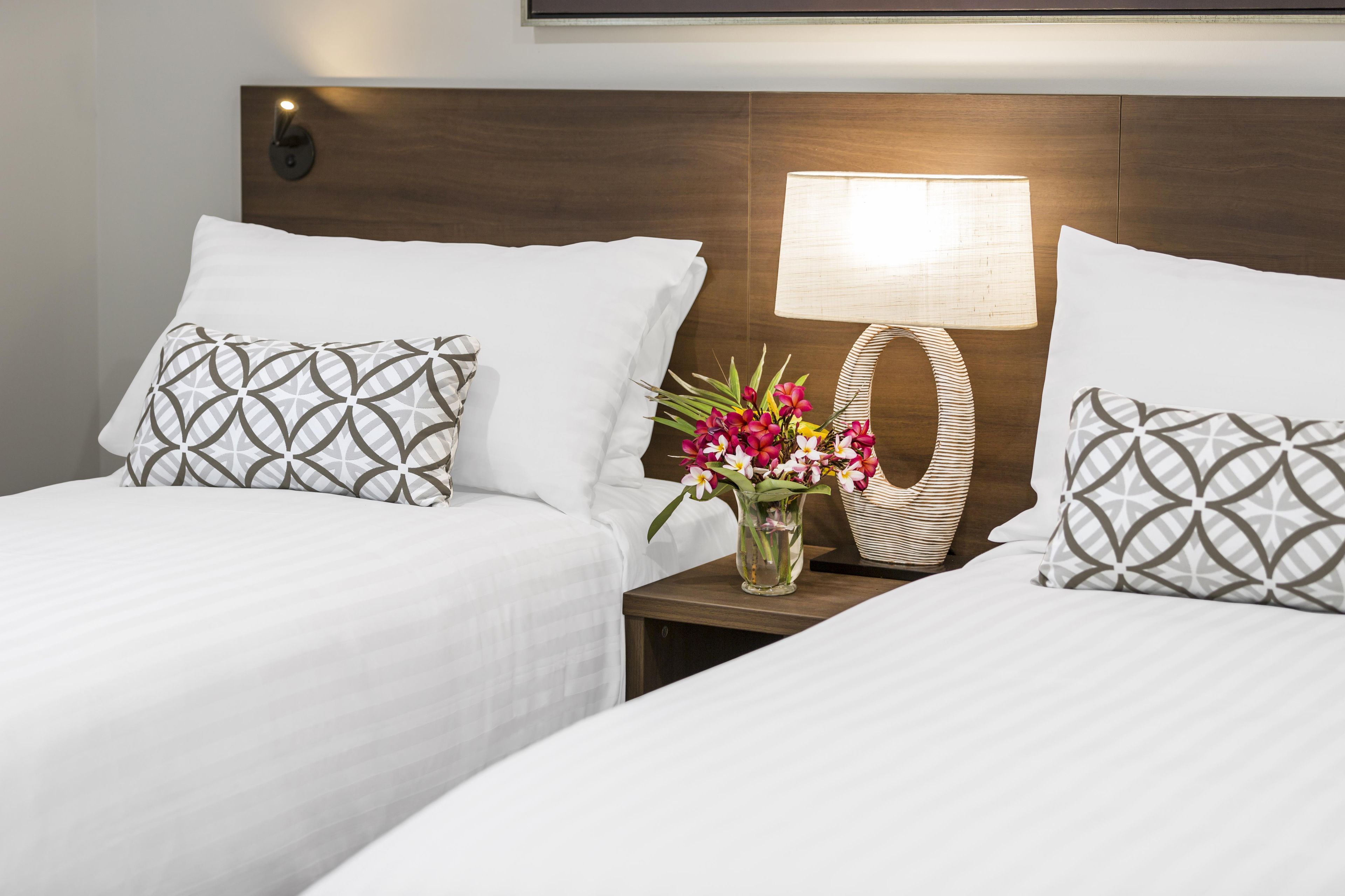 A hotel room with two beds, each with a patterned pillow, a bedside table with a vase of flowers, and a wall-mounted lamp.