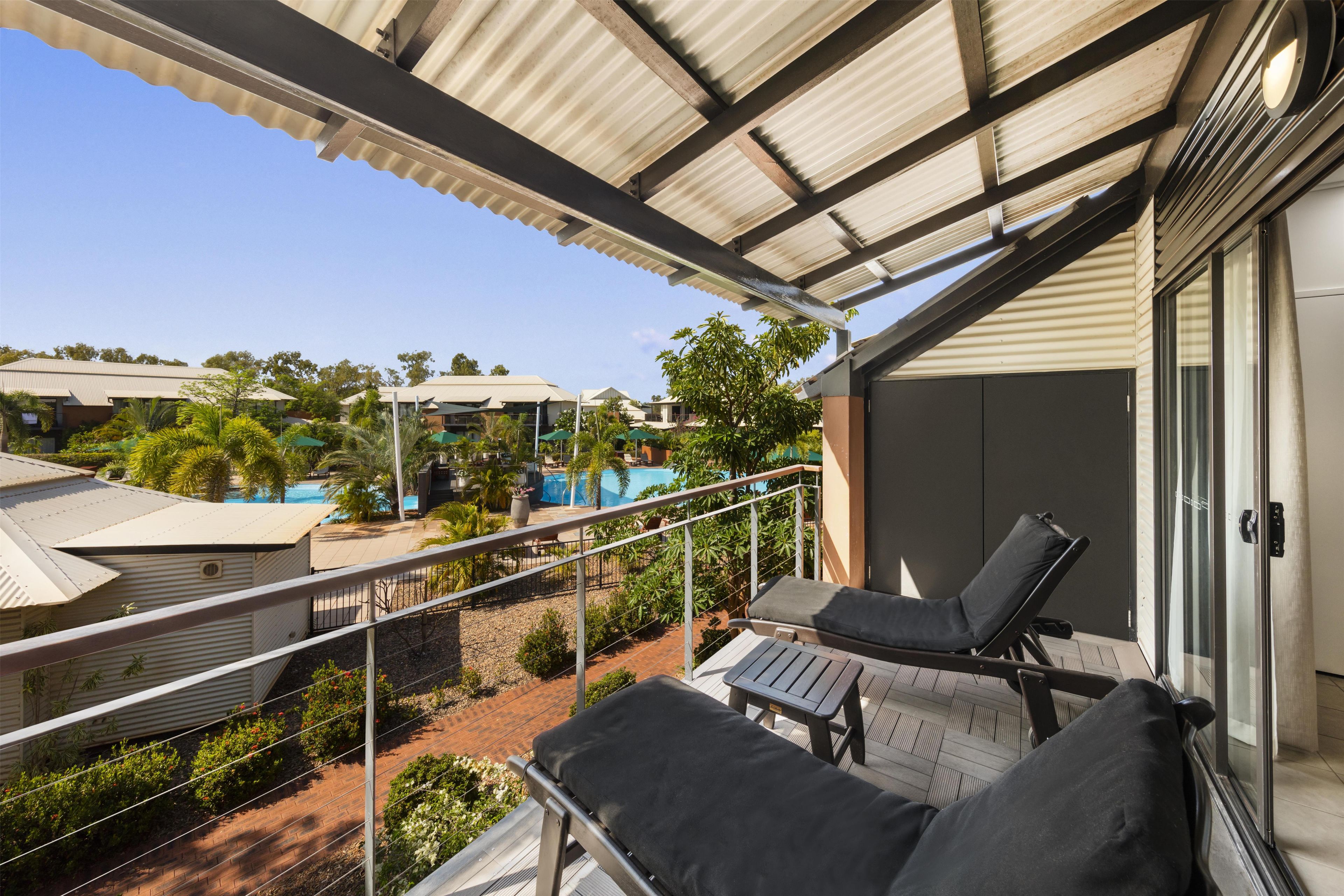 A balcony with two lounge chairs, a view of a swimming pool, and a building with a corrugated metal roof.