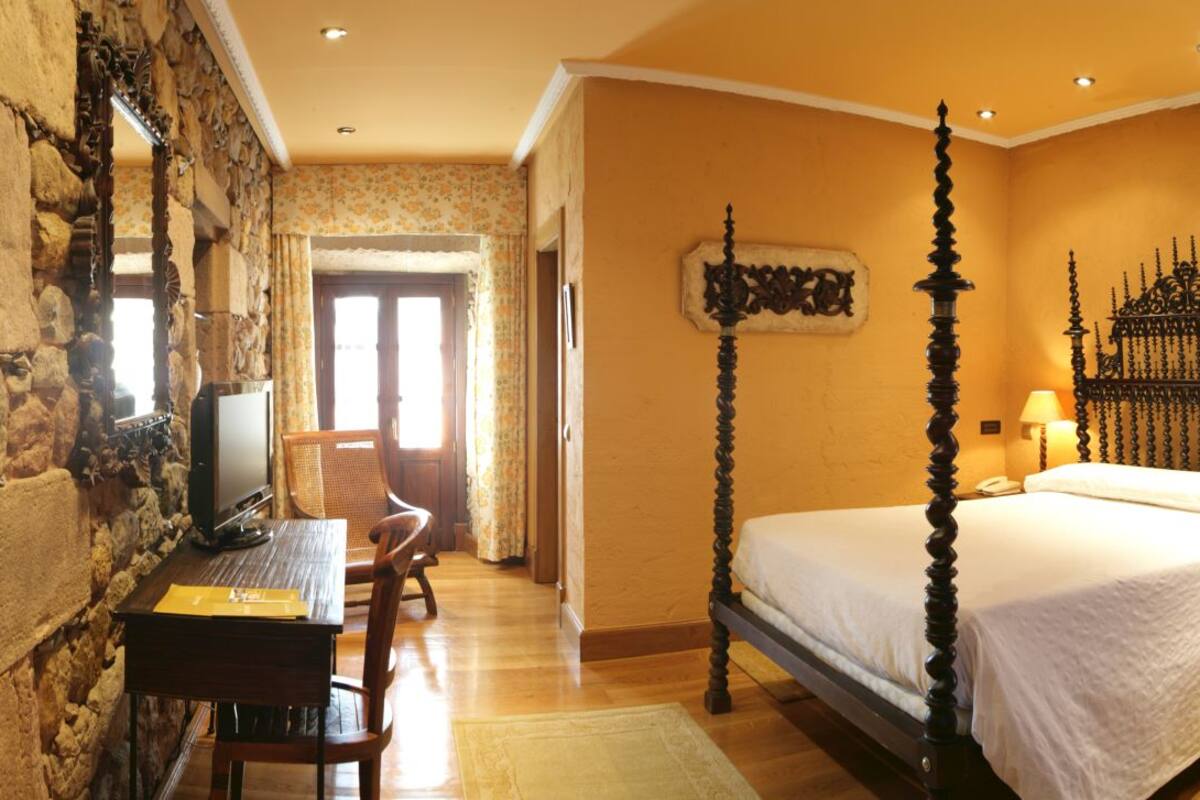 Quarto com cama de dossel, uma escrivaninha com cadeira, parede de pedra e janela com cortinas.