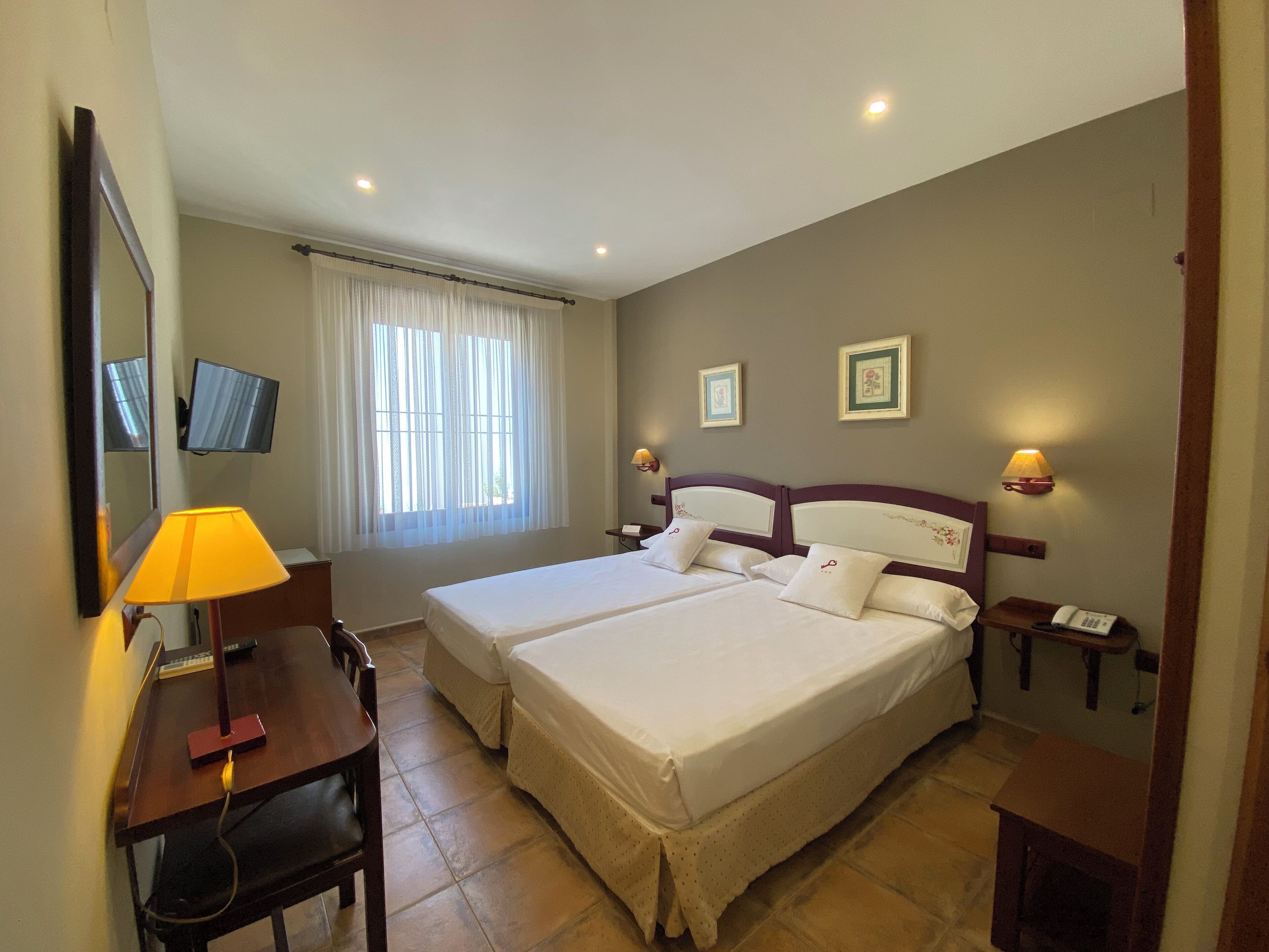 A hotel room with two beds, a desk with a lamp, a TV, and two bedside lamps.