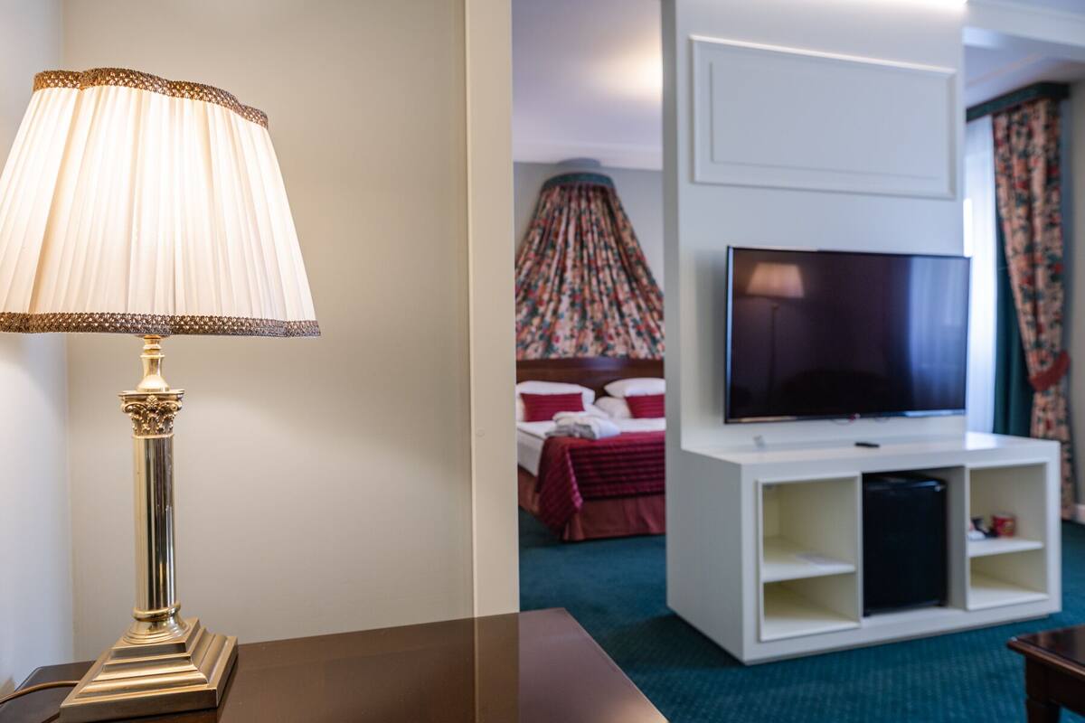 A hotel room with a television on a white stand, a bed with red bedding, and a decorative lamp.