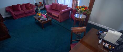 Habitación de hotel con alfombra azul, dos sofás rojos, una mesa de centro de madera, una pequeña mesa redonda con un jarrón de flores y una silla de madera.