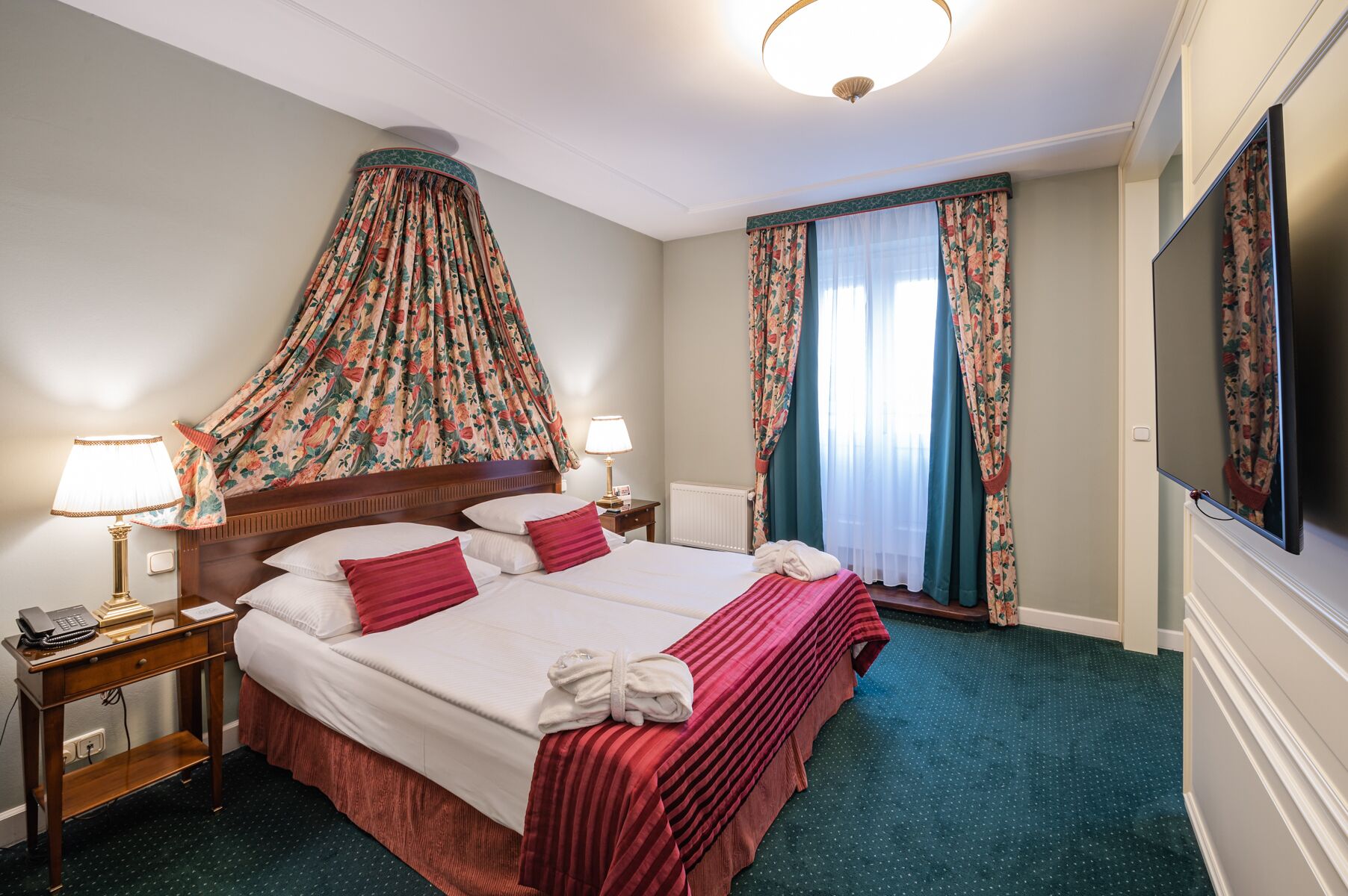 A hotel room with a large bed, two bedside tables with lamps, a television, and a canopy bed with floral curtains.