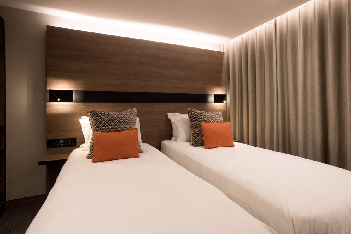 A hotel room with two beds, a wooden headboard, a control panel, and a curtain.