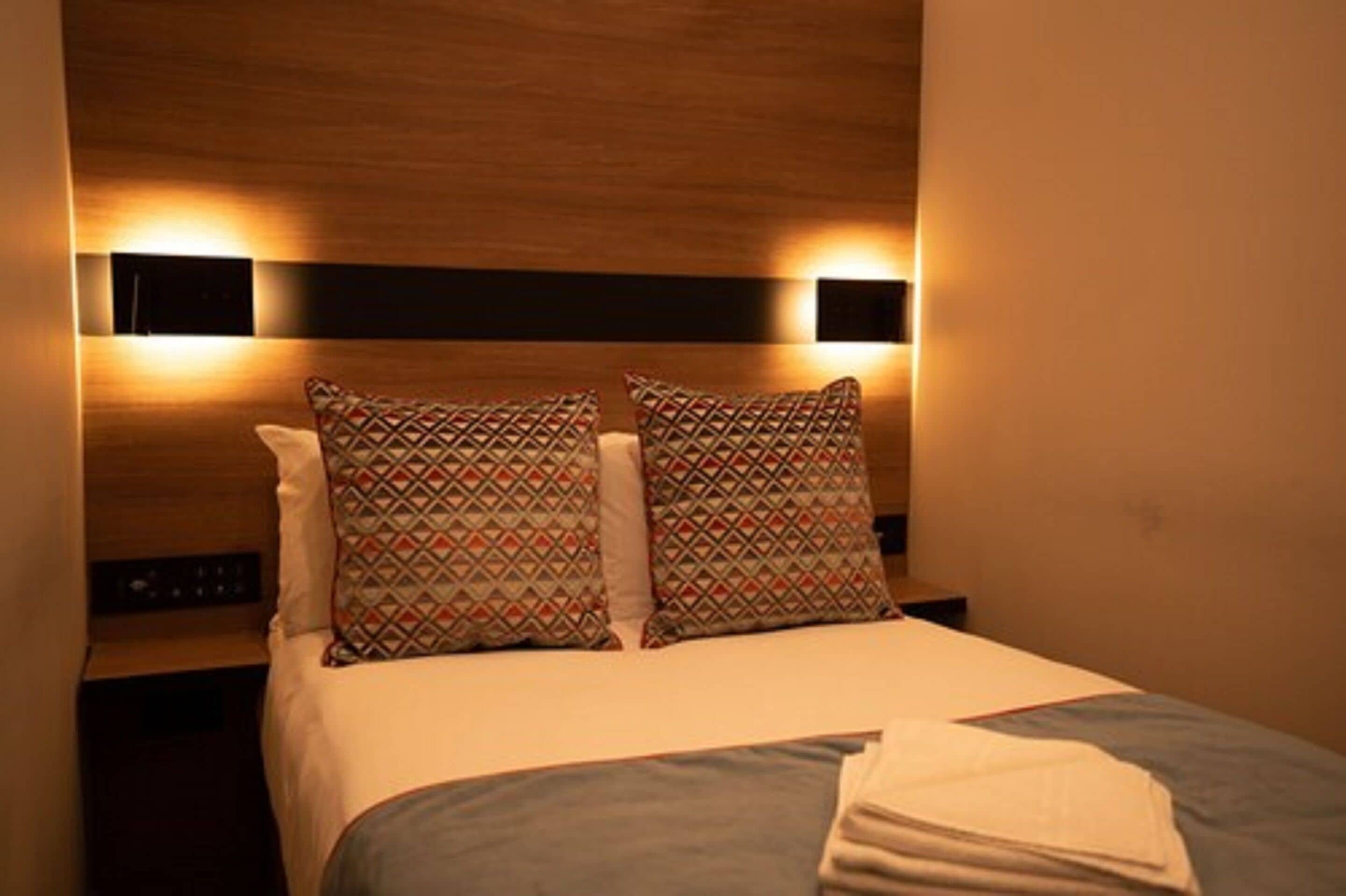 A hotel room with a wooden headboard, two patterned pillows, a white bedspread, and a nightstand with a lamp.