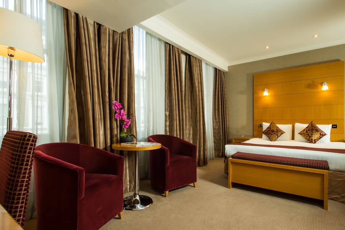 A hotel room with a bed, two red armchairs, a small table with a lamp, and a large window with curtains.