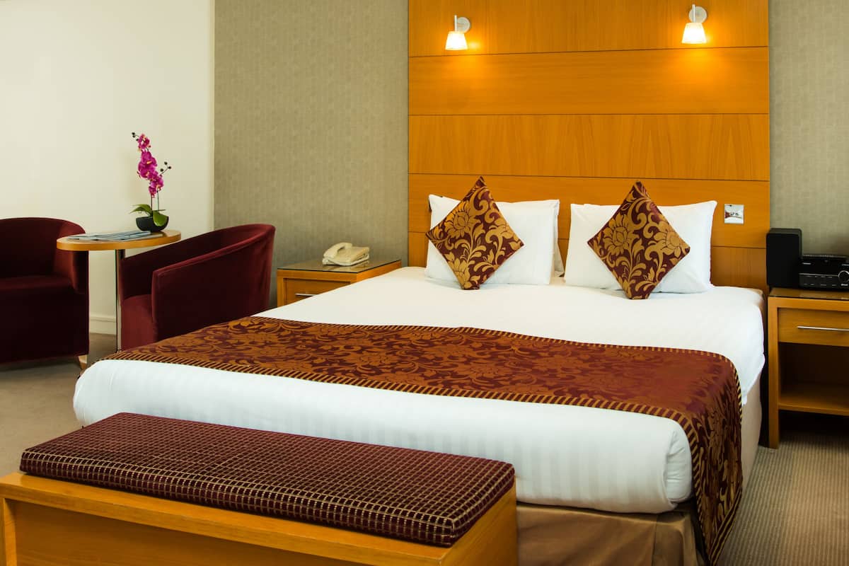 A hotel room with a large bed, a desk with a chair, a small table with a plant, and a bedside table with a phone and a speaker.