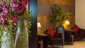 A lobby with large vases of pink flowers, a seating area with brown chairs and red cushions, and a tall plant in the corner. - The Park City Grand Plaza Kensington (London)