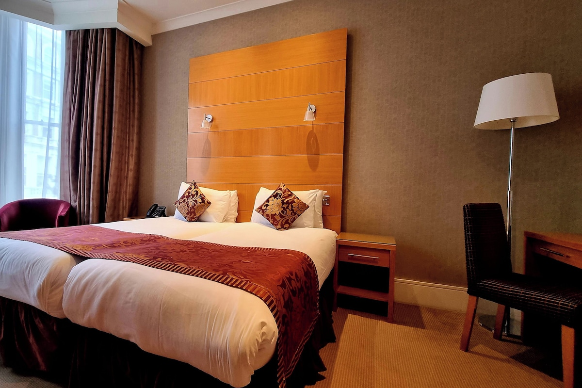 A hotel room with a large bed, a wooden headboard, a desk with a chair, a lamp, and a view of the city through large windows.