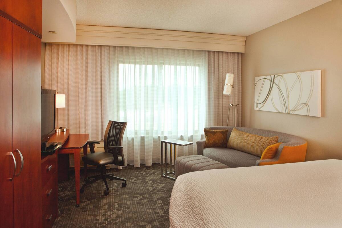 A hotel room with a bed, a desk with a chair, a sofa, a lamp, and a painting on the wall.