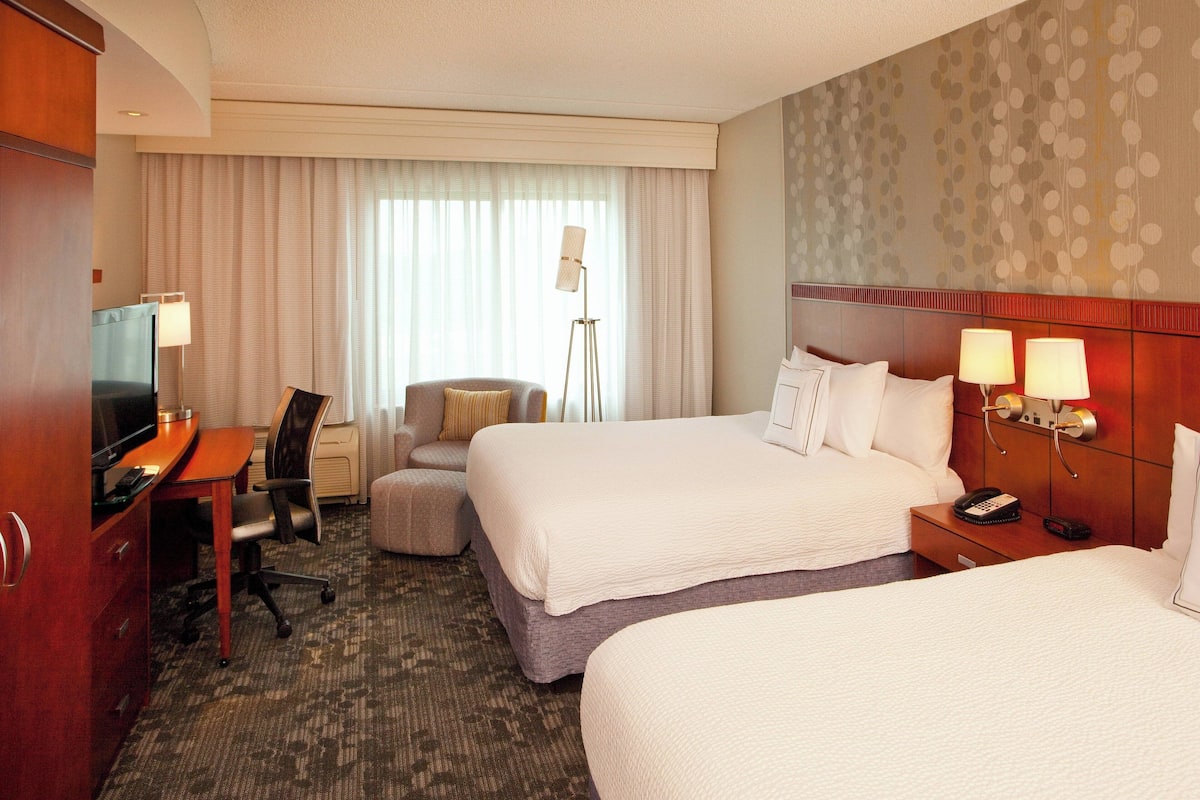 A hotel room with two beds, a desk with a TV, a chair, a lamp, and a bedside table with a phone.