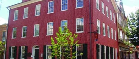 A red brick building with multiple windows, a small tree in front, and a bench.