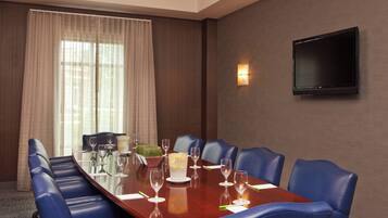 A conference room with a long table, blue chairs, a flat-screen TV, and a window with curtains.