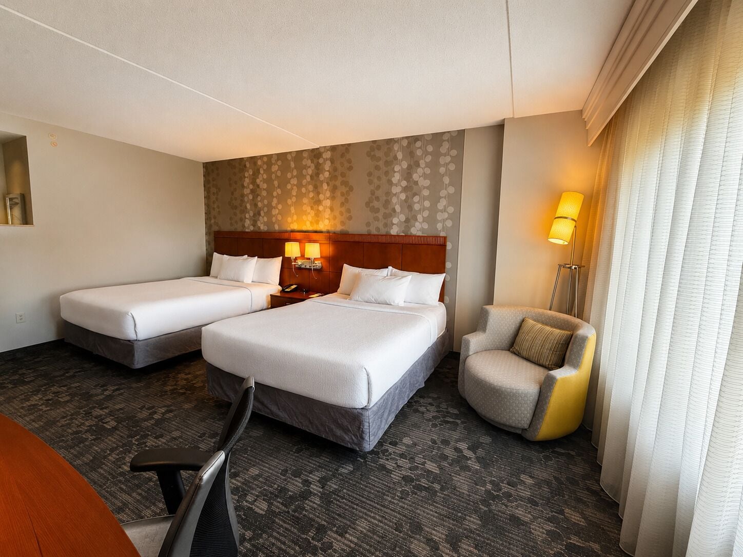 A hotel room with two beds, a desk, a chair, and a lamp.