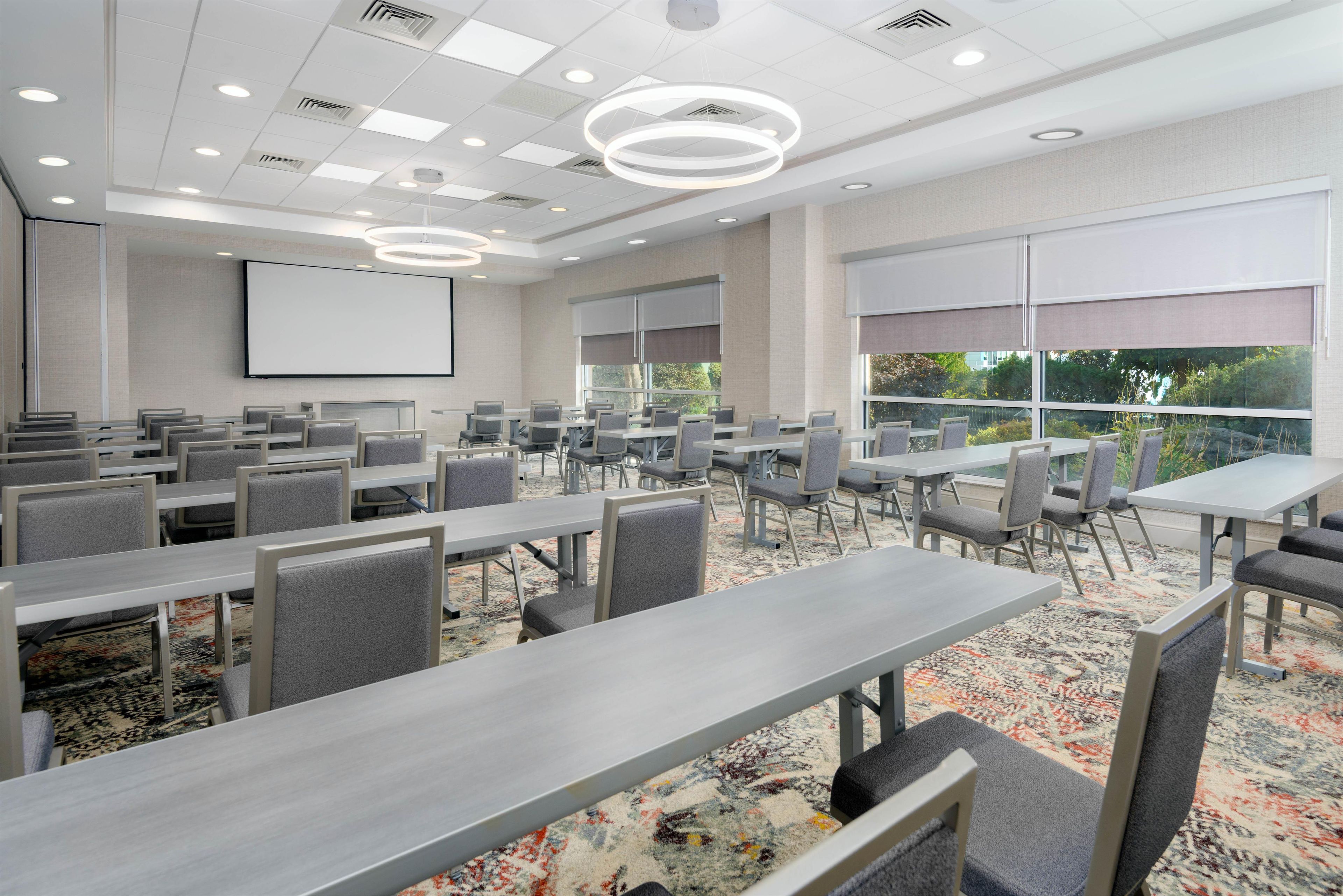 A conference room with multiple tables and chairs, a projector screen, and large windows with blinds.