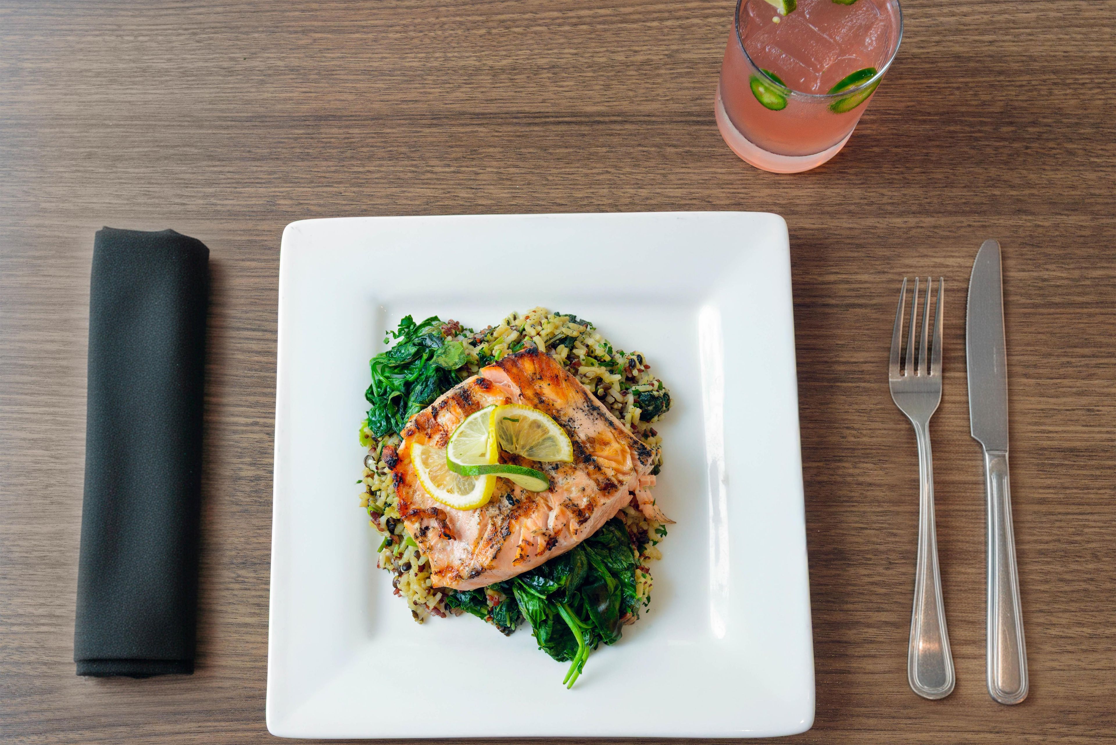 A square white plate with a grilled salmon fillet, spinach, and a grain-based side dish, accompanied by a folded napkin and cutlery on a wooden table.