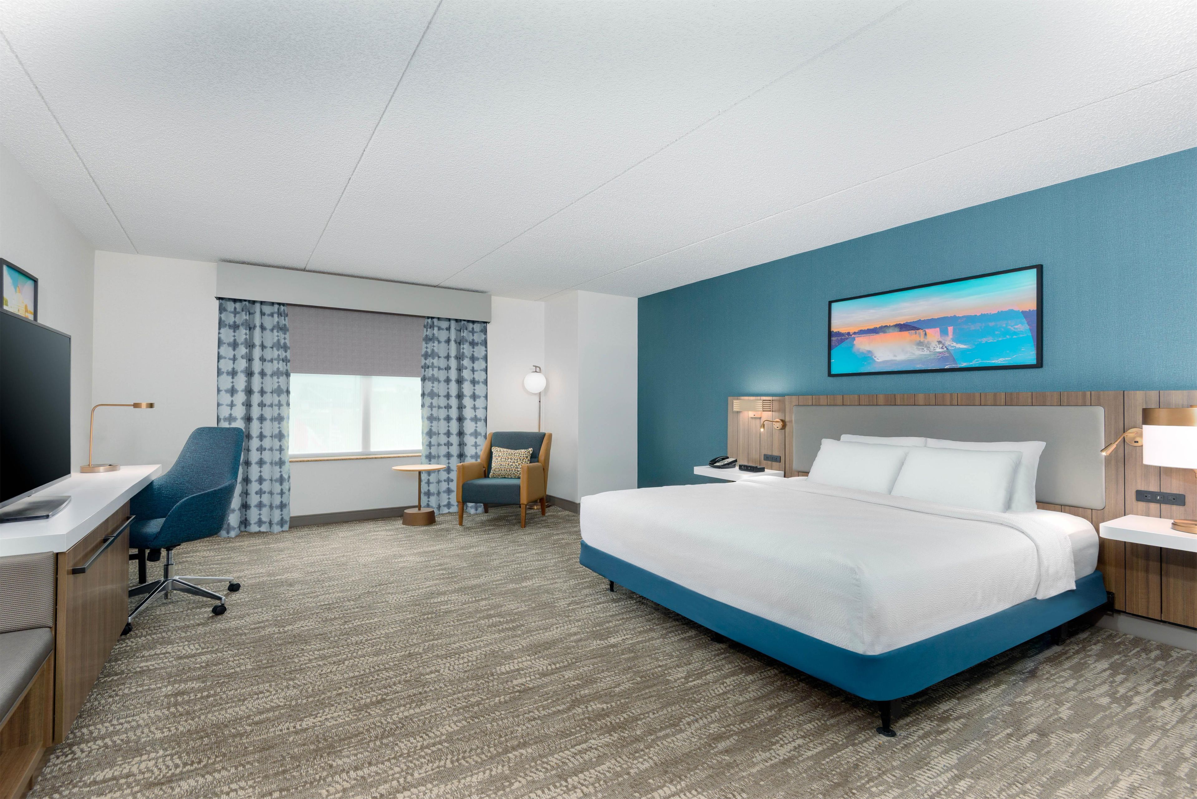 A hotel room with a large bed, a desk with a chair, a small table, a chair, and a painting on the wall.