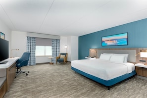 A hotel room with a large bed, a desk with a chair, a small table, a chair, and a painting on the wall. - Hilton Garden Inn Buffalo Airport (Buffalo)