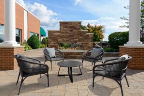 An outdoor patio area with wicker furniture, a stone fire feature, and a brick building in the background. - Hilton Garden Inn Buffalo Airport (Buffalo)