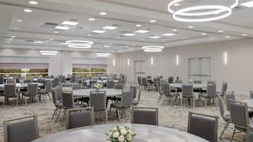 A hotel conference room with multiple tables and chairs, modern lighting fixtures, and a carpeted floor.