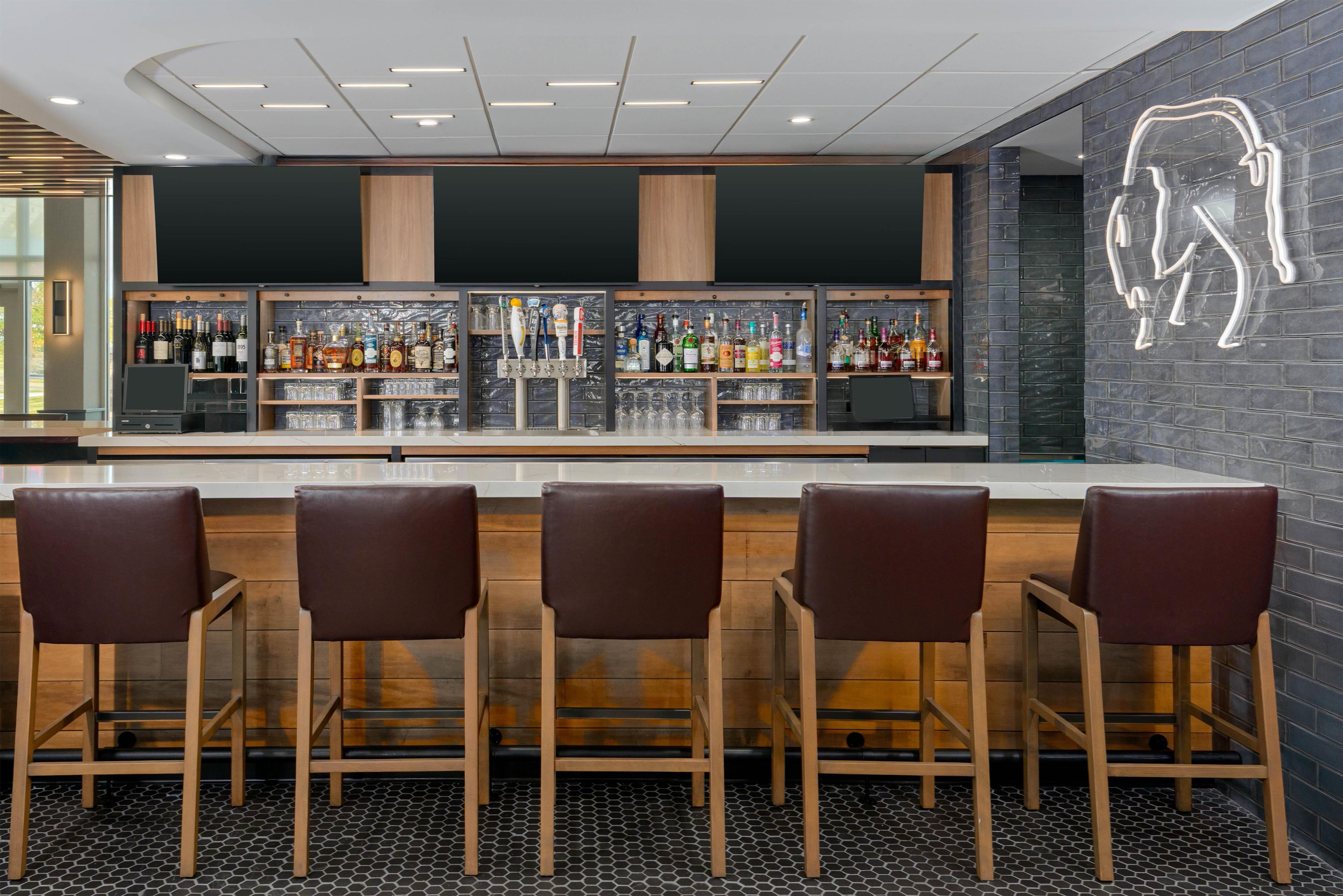 A bar area with a modern design, featuring a counter with multiple taps and shelves of bottles. There are four bar stools in front of the counter.