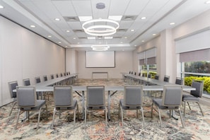 A conference room with a long table and chairs, a projector screen, and a patterned carpet. - Hilton Garden Inn Buffalo Airport (Buffalo)