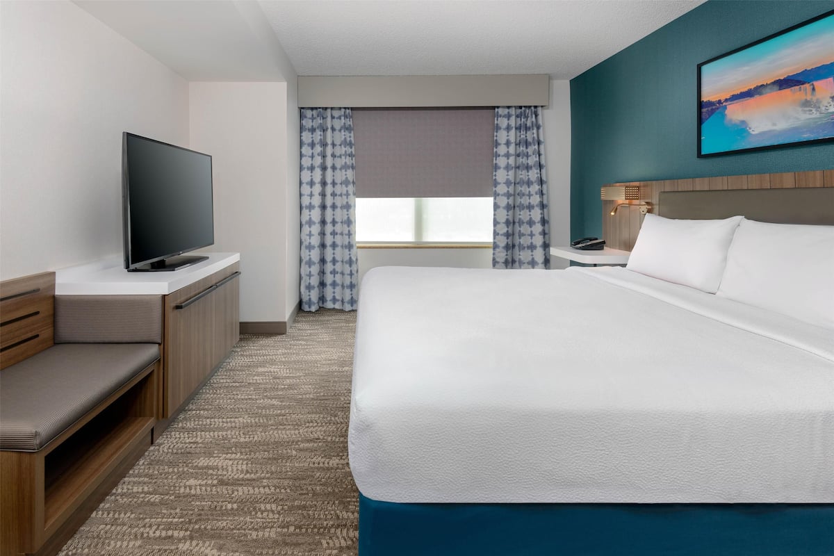 Une chambre d’hôtel avec un grand lit, un téléviseur posé sur un meuble, un bureau avec une lampe et une fenêtre avec des rideaux.