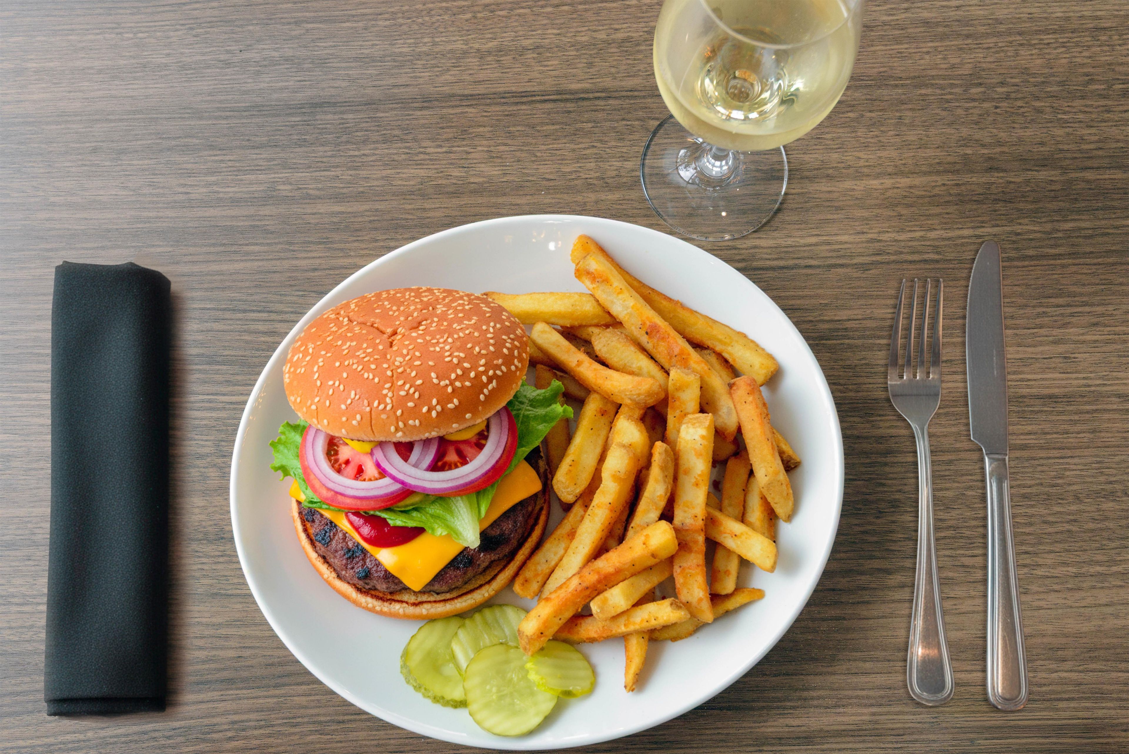 Une assiette contenant un hamburger, des frites et des cornichons, un verre de vin blanc, et des couverts sur une table en bois.