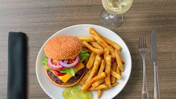 Une assiette contenant un hamburger, des frites et des cornichons, un verre de vin blanc, et des couverts sur une table en bois.