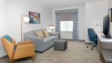 A hotel room with a sofa, armchair, ottoman, desk, television, and a painting on the wall.