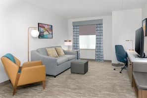 Una habitación de hotel con sofá, sillón, reposapiés, escritorio, televisión y un cuadro en la pared.