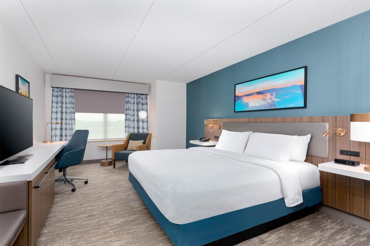 Une chambre d’hôtel avec un grand lit, un bureau, une chaise, une télévision et une fenêtre avec des rideaux.