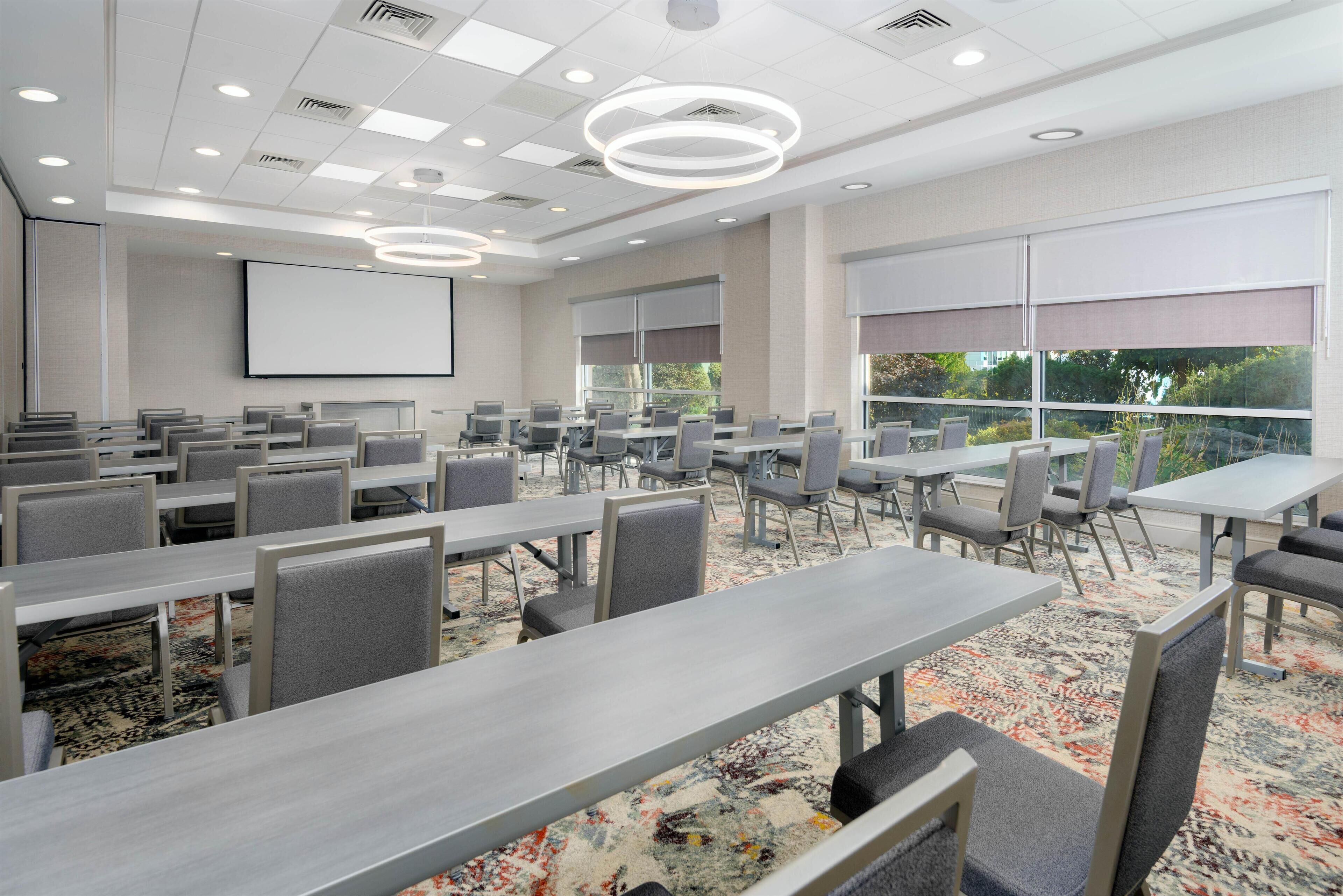 A conference room with multiple tables and chairs, a large screen, and large windows with blinds.