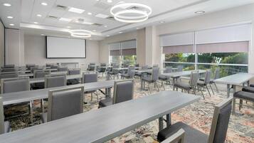 A conference room with multiple tables and chairs, a large screen, and large windows with blinds.