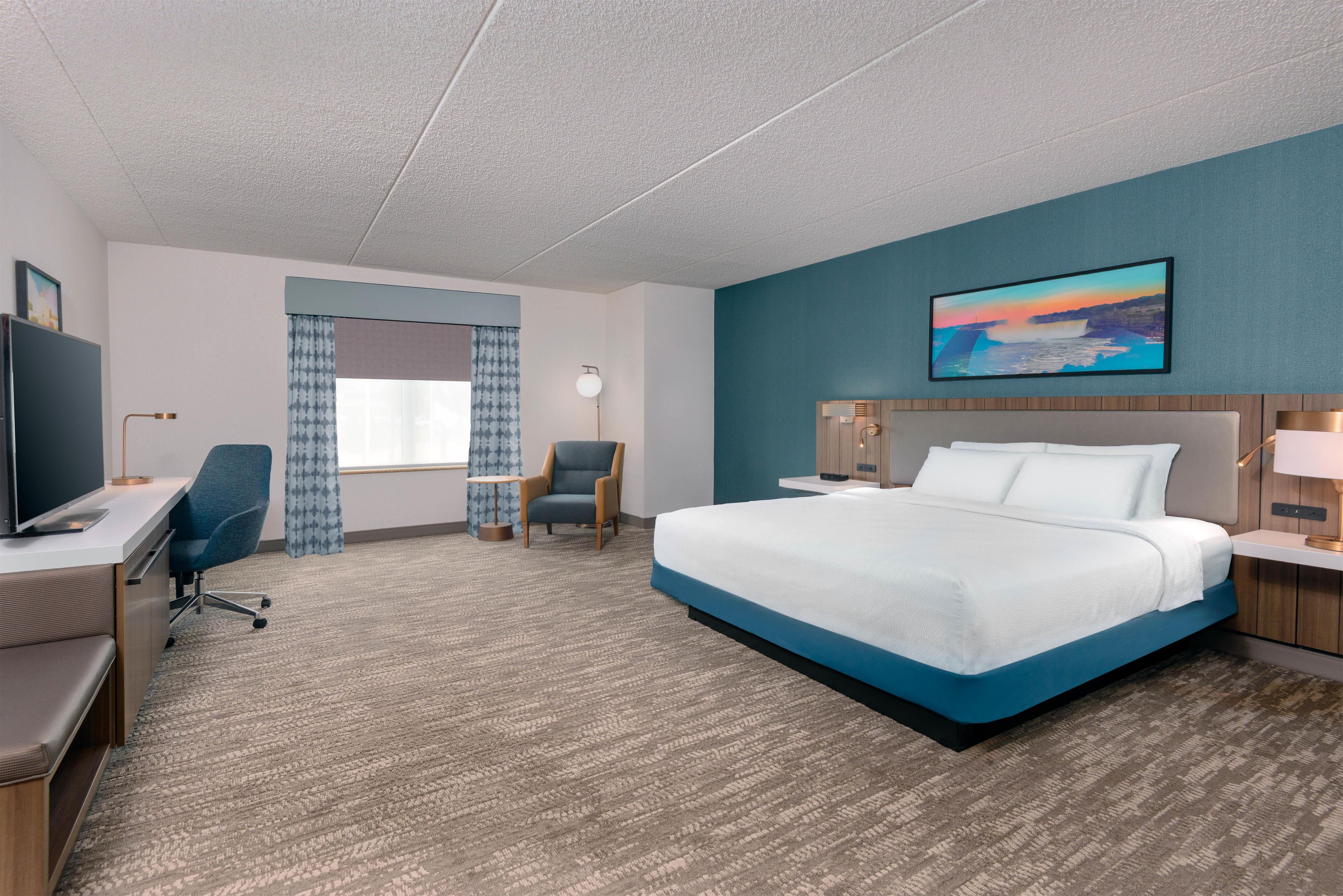 Una habitación de hotel con una cama grande, un escritorio con silla, un televisor, una mesita con silla y un cuadro en la pared.