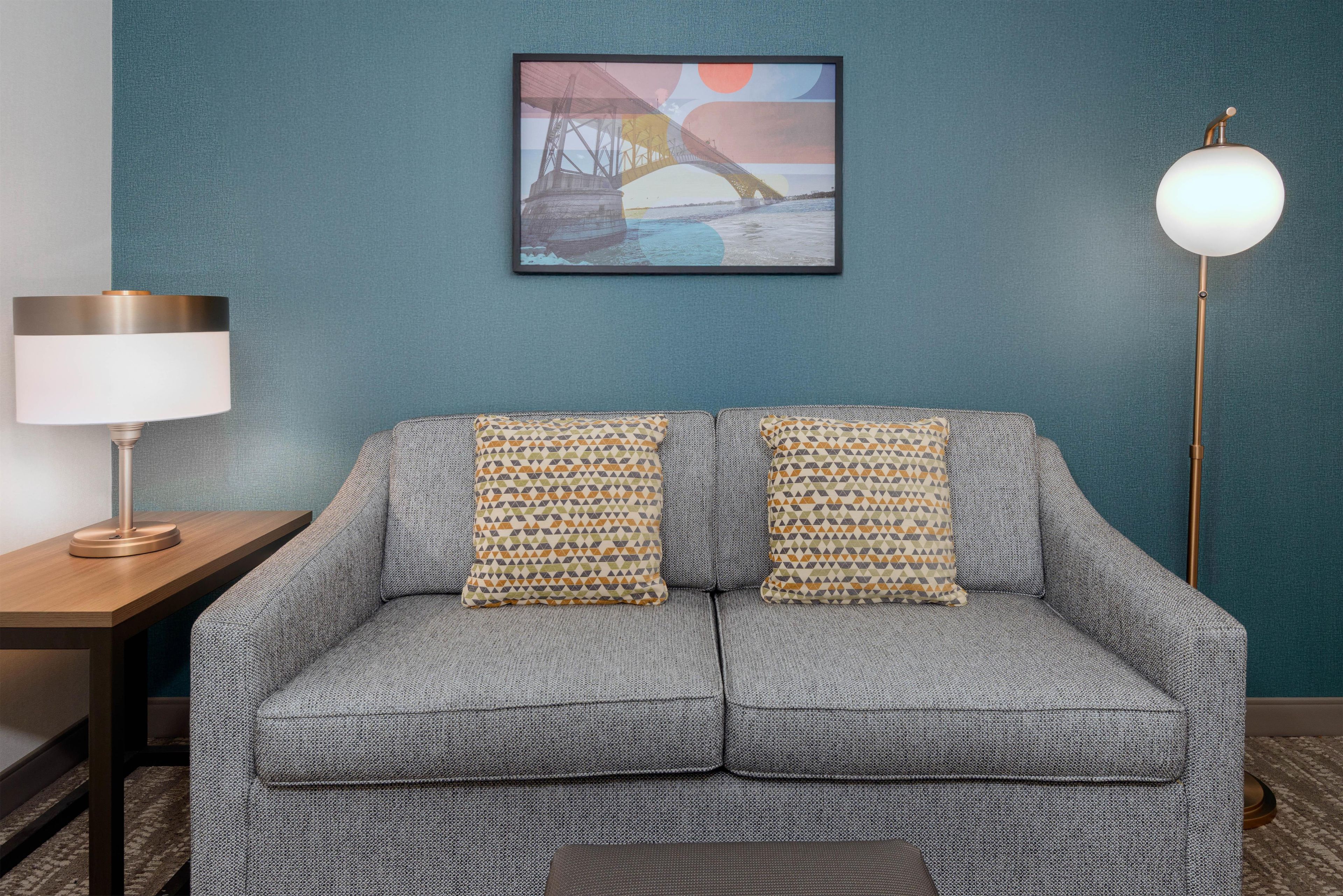 Sofá para dois com almofadas estampadas, mesa lateral com abajur e quadro na parede.
