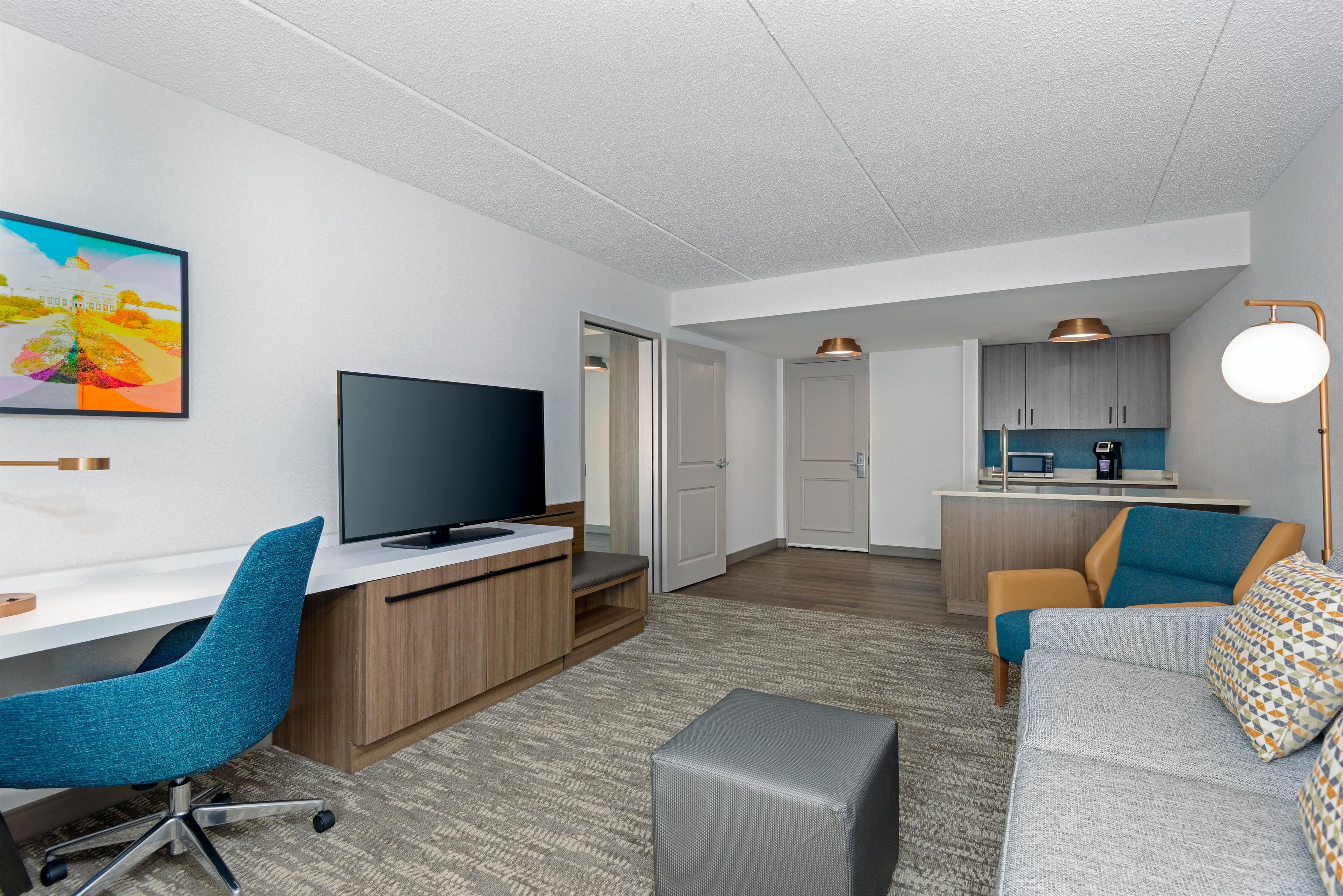 Quarto de hotel moderno com televisão de tela plana, escrivaninha, cadeira azul, sofá cinza e área de cozinha com micro-ondas.