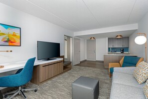 Une chambre d’hôtel moderne équipée d’un téléviseur à écran plat, d’un bureau, d’une chaise bleue, d’un canapé gris et d’un coin cuisine avec un four à micro-ondes.