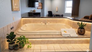 A bathroom with a large bathtub, a chair, a television, and a window with curtains. - Apple Tree Inn and Suites (Independence)