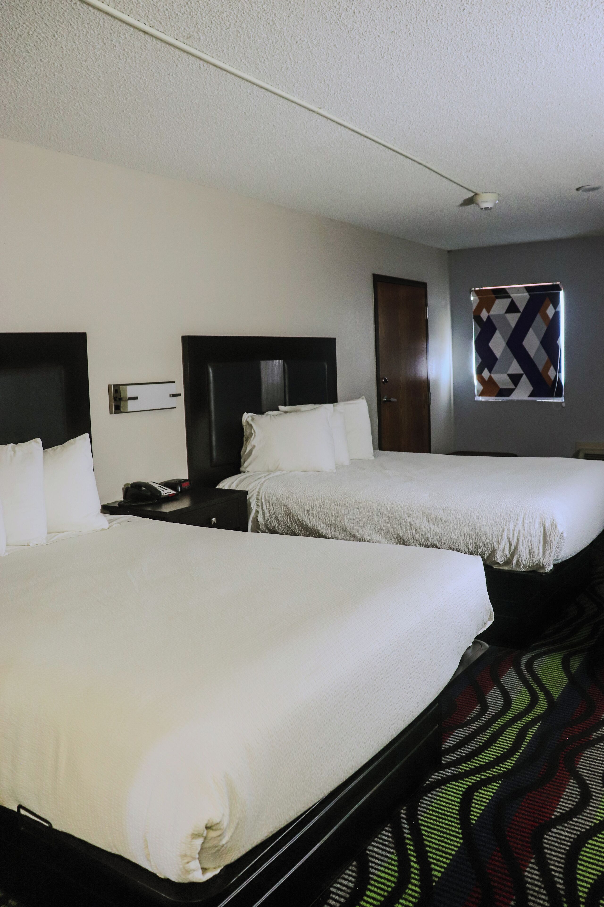 A hotel room with two beds, a nightstand with a phone, a wall-mounted TV, and a window with a geometric curtain.