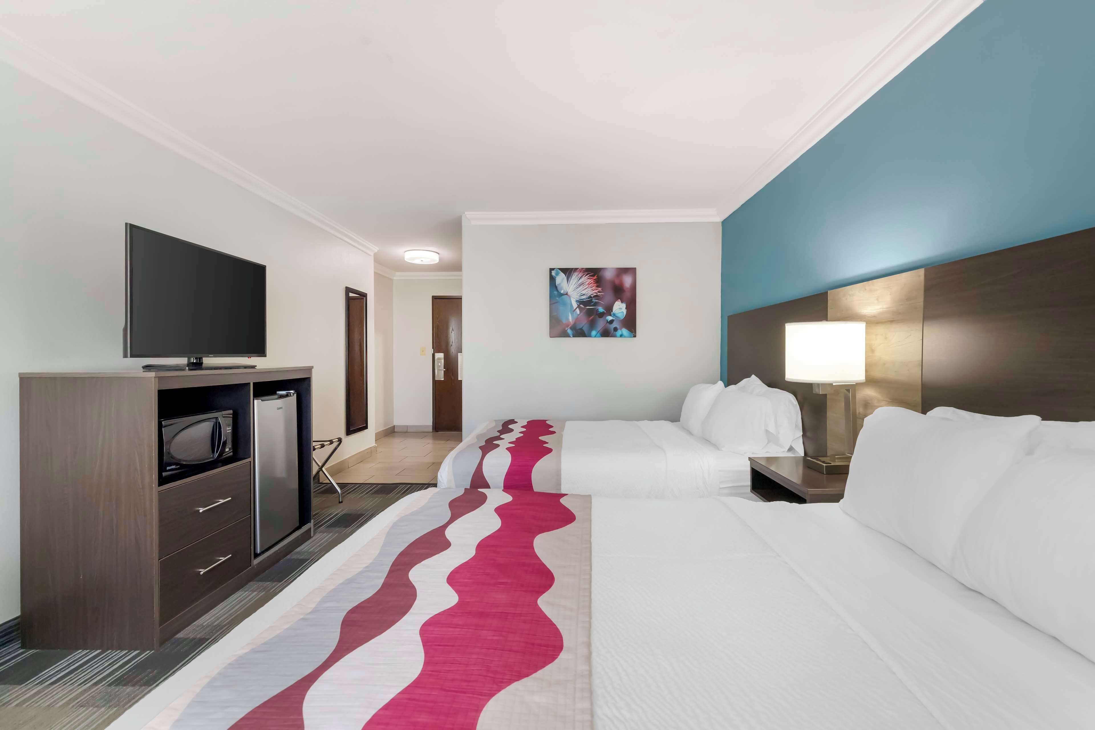 A hotel room with two beds, a flat-screen TV, a minibar, and a painting on the wall.